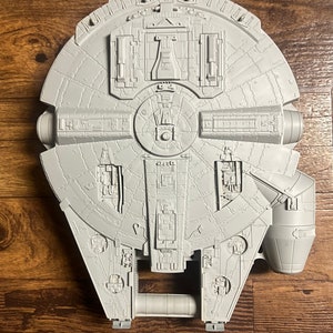 May include: A gray plastic model of the Millennium Falcon spaceship from Star Wars. The model is detailed with various hatches, panels, and other features.
