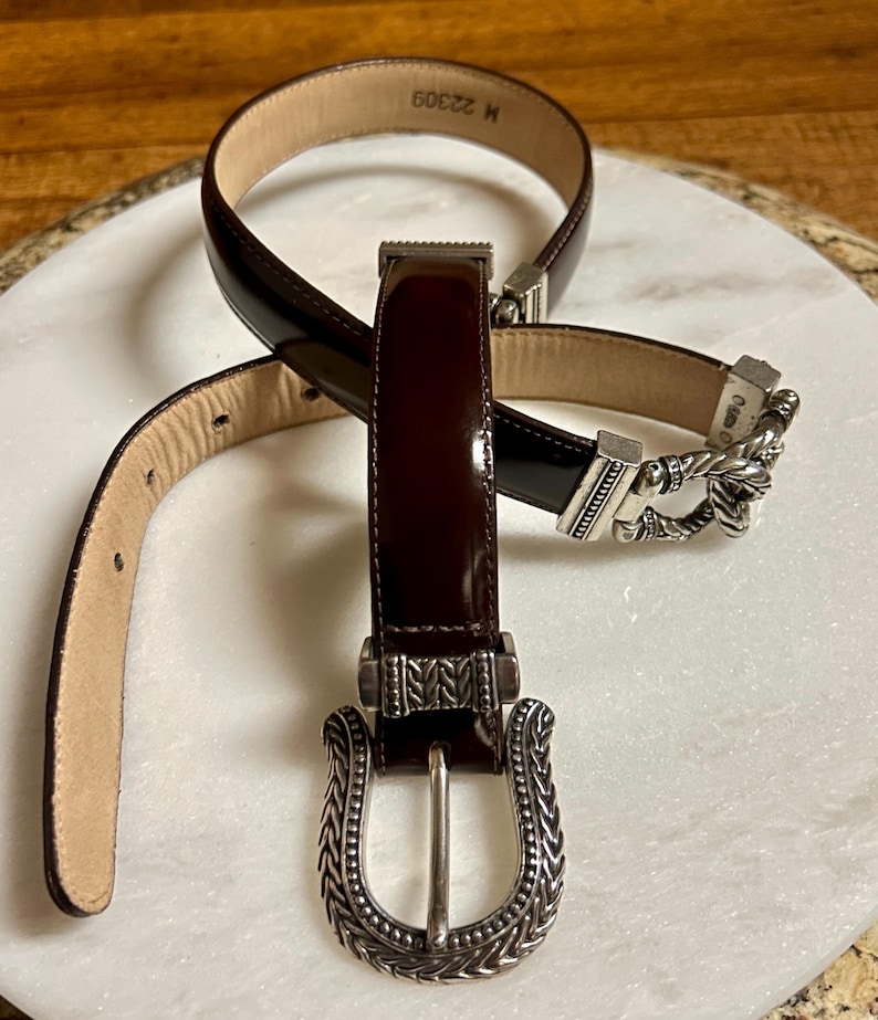 Puede incluir: Dos cinturones de cuero marr&oacute;n con hebillas plateadas. La hebilla del cintur&oacute;n superior tiene un dise&ntilde;o trenzado y la hebilla del cintur&oacute;n inferior tiene un dise&ntilde;o de cuerda. El cintur&oacute;n superior tiene el texto "M 32309" en la parte posterior.