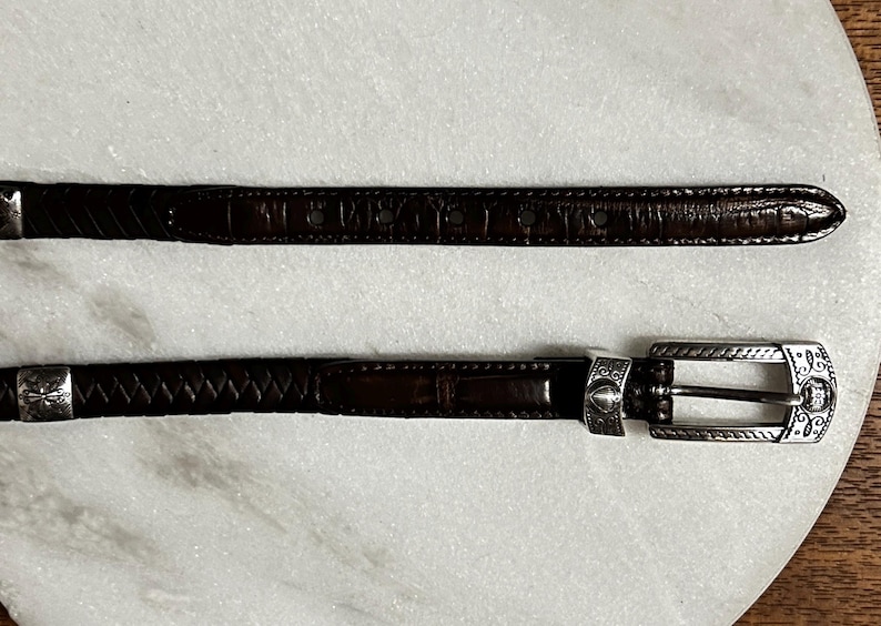 K&ouml;nnte beinhalten: Zwei braune Lederg&uuml;rtel mit silberfarbenen Schnallen. Einer hat eine strukturierte Krokodil-Optik, der andere ein geflochtenes Design. Die Schnallen sind mit dekorativen Details versehen.