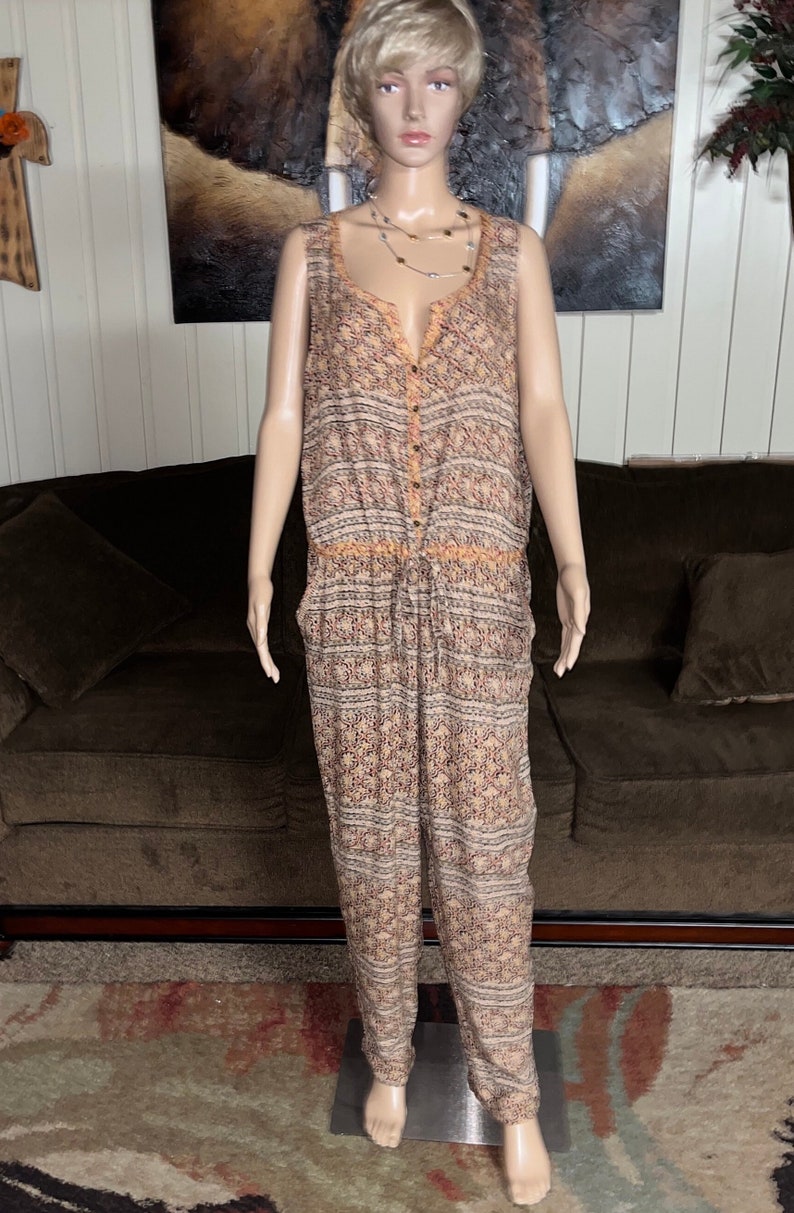 May include: A sleeveless, button-down jumpsuit with a geometric pattern in shades of brown, orange, and white. The jumpsuit has a drawstring waist and wide legs.