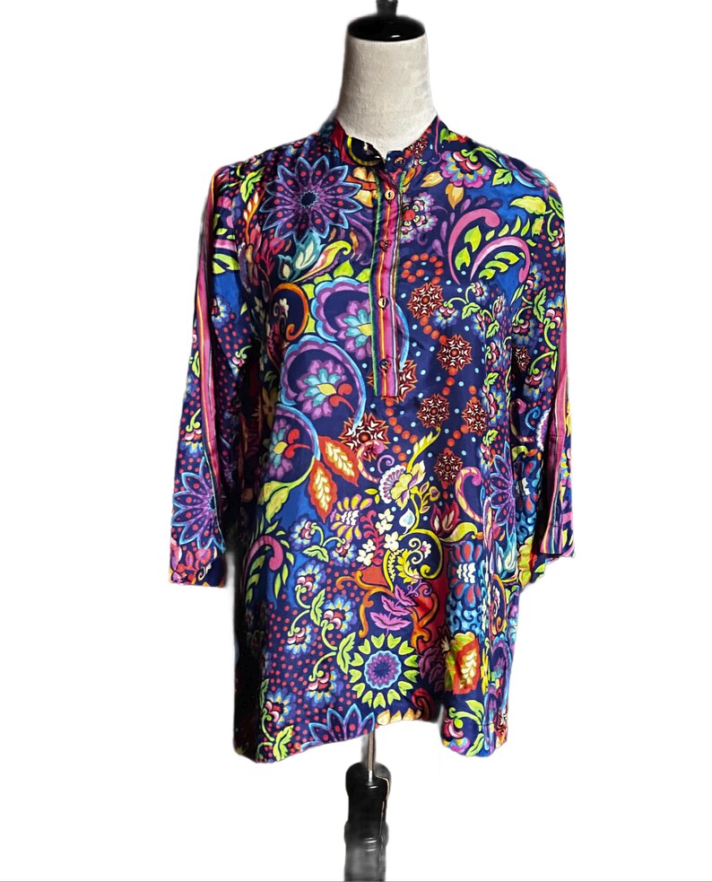 May include: A long-sleeved tunic with a vibrant floral and paisley pattern in shades of blue, purple, pink, yellow, and green. The tunic has a mandarin collar and a button-down placket. The sleeves are three-quarter length.