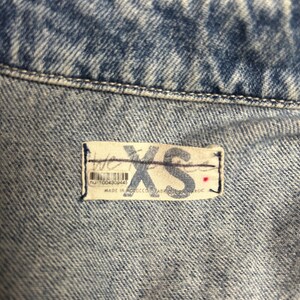 Peut inclure: Gros plan sur un tissu en jean bleu clair avec une &eacute;tiquette blanche indiquant "XS" et "nu 1004309445" avec un point rouge. L'&eacute;tiquette est cousue sur le tissu.