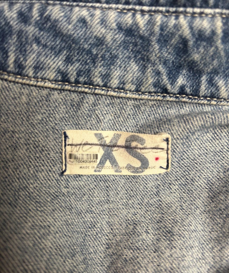Peut inclure: Gros plan sur un tissu en jean bleu clair avec une &eacute;tiquette blanche indiquant "XS" et "nu 1004309445" avec un point rouge. L'&eacute;tiquette est cousue sur le tissu.