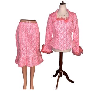 May include: A two-piece pink lace suit, consisting of a fitted jacket with bell sleeves and a pencil skirt with a ruffled hem. The jacket is buttoned and displayed on a mannequin. The skirt is displayed on a mannequin.