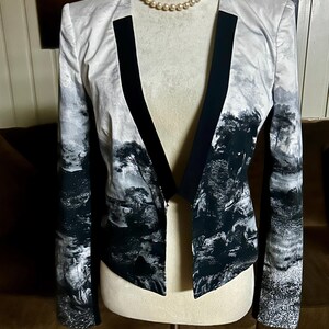 May include: A black and white landscape print blazer with a black lapel and trim. The open-front jacket is displayed on a mannequin with a pearl necklace. The landscape design features trees and water elements.