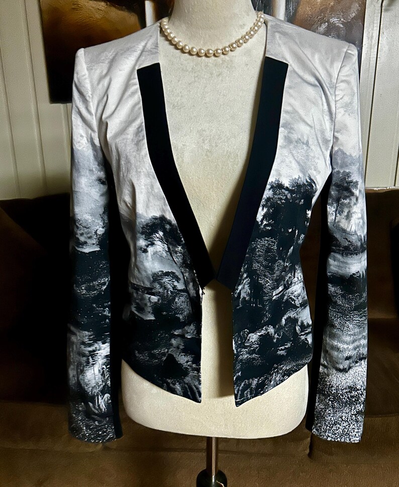 May include: A black and white landscape print blazer with a black lapel and trim. The open-front jacket is displayed on a mannequin with a pearl necklace. The landscape design features trees and water elements.