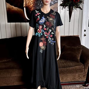 Puede incluir: Un vestido negro de largo midi con mangas cortas y cuello redondo. La parte delantera del vestido presenta bordados coloridos que representan un p&aacute;jaro azul, varias flores en tonos rojos, naranjas y amarillos, y peque&ntilde;os motivos de fresas.