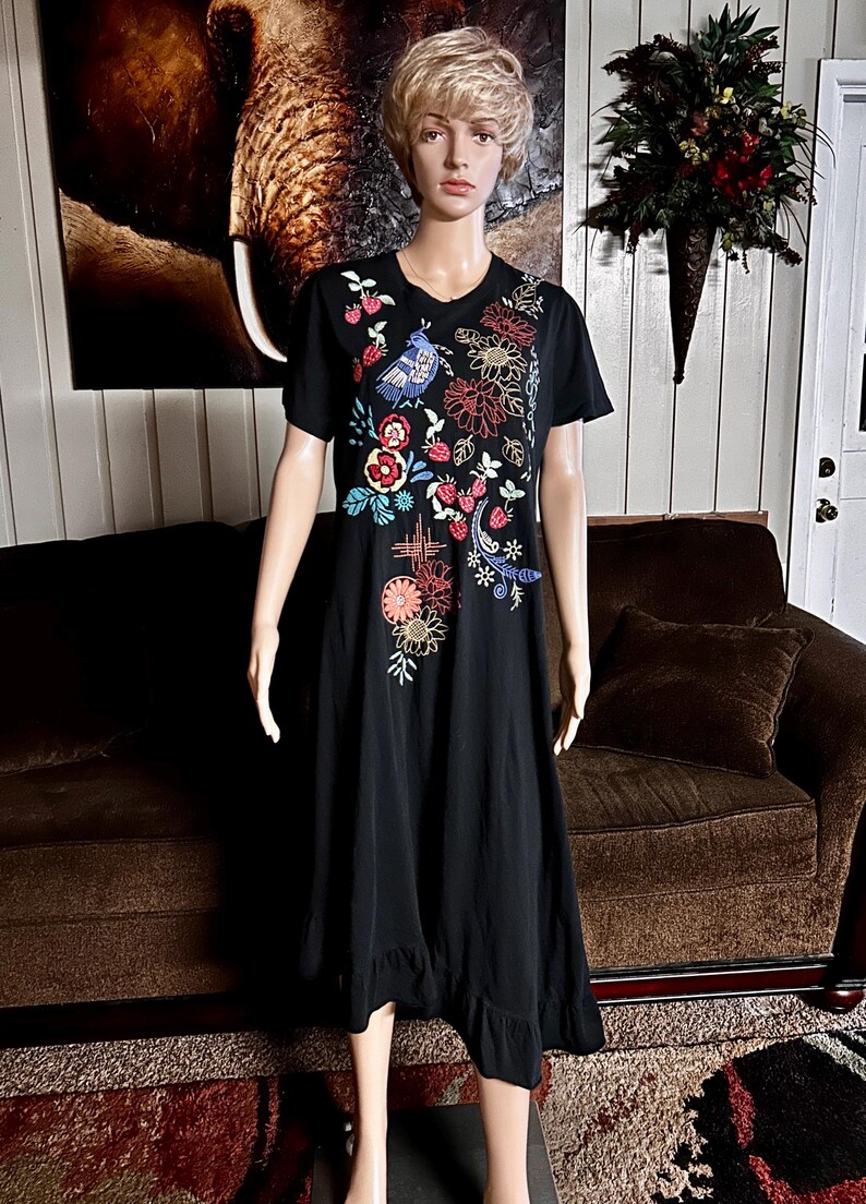 Puede incluir: Un vestido negro de largo midi con mangas cortas y cuello redondo. La parte delantera del vestido presenta bordados coloridos que representan un p&aacute;jaro azul, varias flores en tonos rojos, naranjas y amarillos, y peque&ntilde;os motivos de fresas.