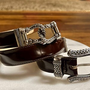 Puede incluir: Un cintur&oacute;n de cuero marr&oacute;n con una hebilla plateada. La hebilla tiene un dise&ntilde;o trenzado y un cierre decorativo.