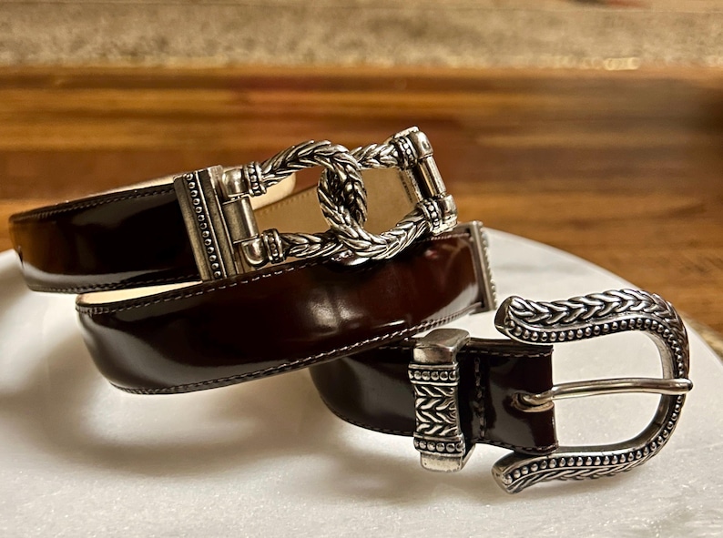 Puede incluir: Un cintur&oacute;n de cuero marr&oacute;n con una hebilla plateada. La hebilla tiene un dise&ntilde;o trenzado y un cierre decorativo.