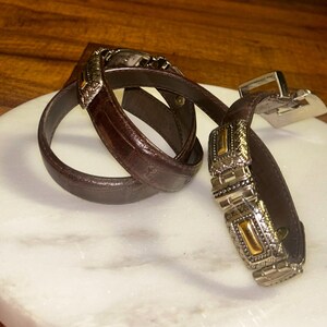 May include: A brown leather belt with ornate silver and gold-toned buckle and accents. The belt is coiled on a white marble surface, showcasing its intricate design and craftsmanship. The buckle features detailed patterns.