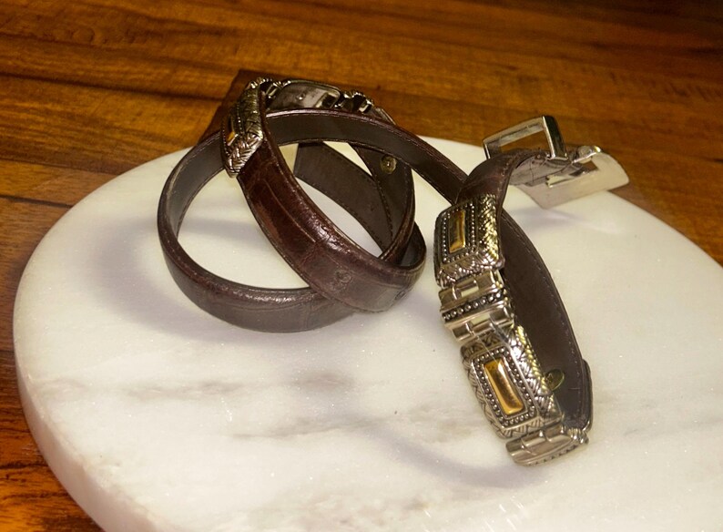 May include: A brown leather belt with ornate silver and gold-toned buckle and accents. The belt is coiled on a white marble surface, showcasing its intricate design and craftsmanship. The buckle features detailed patterns.