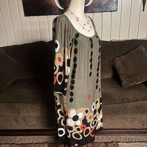 May include: A green, black, white, and orange patterned dress with a scoop neck and 3/4 sleeves. The dress is on a mannequin and is displayed against a brown couch.