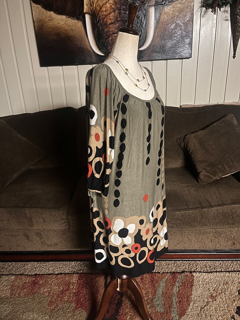 May include: A green, black, white, and orange patterned dress with a scoop neck and 3/4 sleeves. The dress is on a mannequin and is displayed against a brown couch.