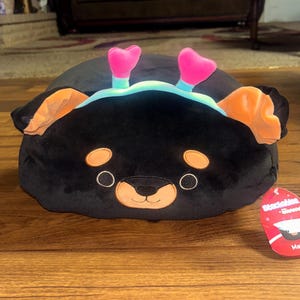 May include: A black plush toy shaped like a bear with orange ears and pink heart-shaped feet. The bear has a tan snout and a tag that says "Mateo". The toy is on a wooden surface.