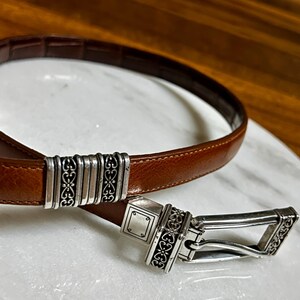 May include: A brown leather belt with a silver-toned buckle and decorative accents. The belt features a rectangular buckle with ornate scrollwork and a matching keeper. The leather is a rich, warm brown.