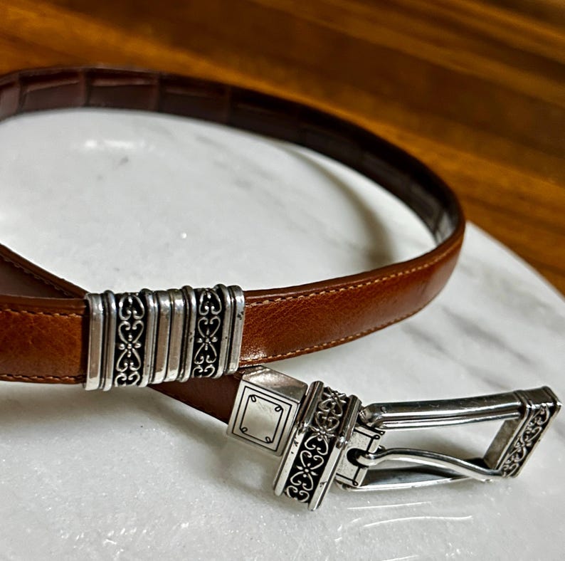 May include: A brown leather belt with a silver-toned buckle and decorative accents. The belt features a rectangular buckle with ornate scrollwork and a matching keeper. The leather is a rich, warm brown.