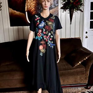 Puede incluir: Un vestido midi negro de manga corta con un vibrante patr&oacute;n de bordado floral y de aves en varios colores en la parte superior delantera. El dise&ntilde;o incluye hilos rojos, amarillos, azules y verdes que representan flores, hojas y un ave.