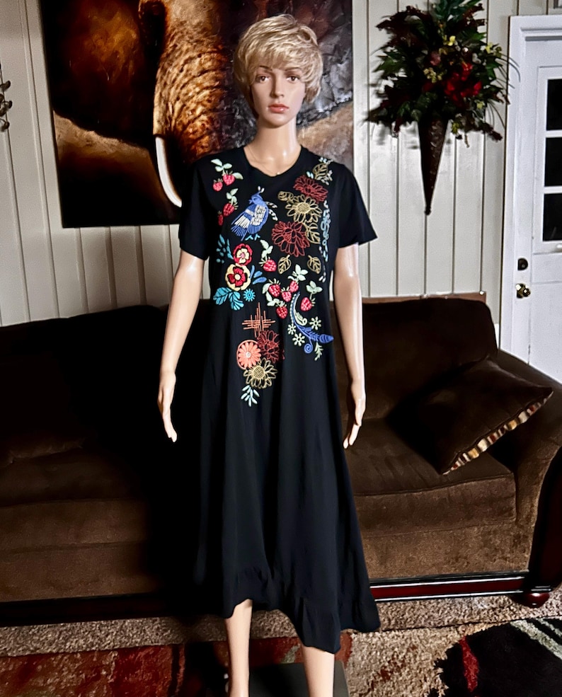 Puede incluir: Un vestido midi negro de manga corta con un vibrante patr&oacute;n de bordado floral y de aves en varios colores en la parte superior delantera. El dise&ntilde;o incluye hilos rojos, amarillos, azules y verdes que representan flores, hojas y un ave.