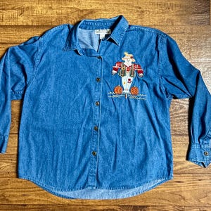 May include: A blue denim button-down shirt with long sleeves. The shirt features an embroidered scarecrow design with pumpkins. The scarecrow is wearing a hat, overalls, and a plaid shirt. The shirt has a collared neckline and button closures.