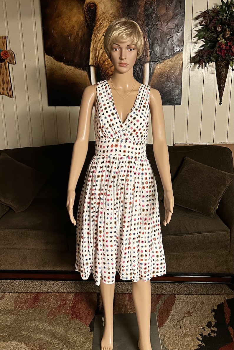 May include: White sleeveless dress with a V-neck and a full skirt. The dress is covered in a pattern of colorful polka dots. The dress is displayed on a mannequin.