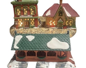 May include: A Christmas village scene featuring a red brick building with the text "Woodbridge Post Office", a house with a red roof, and a green-roofed bridge. The buildings are covered in snow and lit with warm lights.