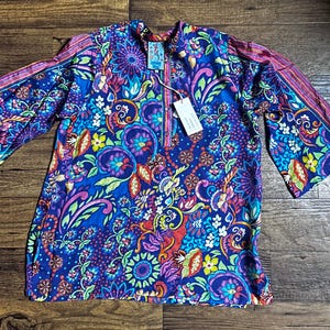 May include: A vibrant, long-sleeved tunic with a colorful floral and paisley pattern in shades of blue, purple, pink, and green. The tunic has a mandarin collar and a zipper closure. The sleeves have a contrasting striped design.