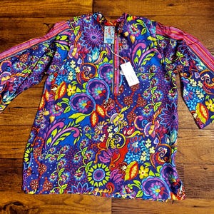 May include: A vibrant, patterned tunic in shades of blue, purple, pink, and yellow. The tunic features a floral design with bell sleeves and a mandarin collar. A tag is attached to the tunic.