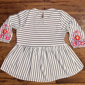 May include: White and black striped blouse with a ruffled hem and floral embroidery on the sleeves.