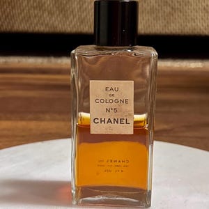 May include: A rectangular glass bottle of Chanel No. 5 Eau de Cologne. The bottle has a black cap and a beige label with black text. The liquid inside is amber-colored. The bottle is sitting on a white surface.