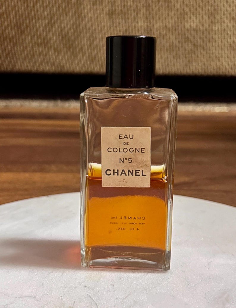 May include: A rectangular glass bottle of Chanel No. 5 Eau de Cologne. The bottle has a black cap and a beige label with black text. The liquid inside is amber-colored. The bottle is sitting on a white surface.