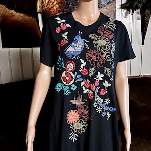 Puede incluir: Una camiseta negra de manga corta con coloridos bordados de un p&aacute;jaro azul, fresas rojas y varias flores en tonos rojos, naranjas, amarillos y azules. El bordado cubre la parte delantera de la prenda con un patr&oacute;n disperso.