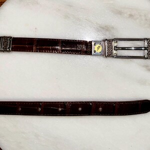 May include: A brown leather belt with a crocodile texture. The belt has a silver buckle with decorative detailing and a silver loop. The belt is laid out on a white surface.