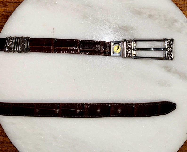 May include: A brown leather belt with a crocodile texture. The belt has a silver buckle with decorative detailing and a silver loop. The belt is laid out on a white surface.
