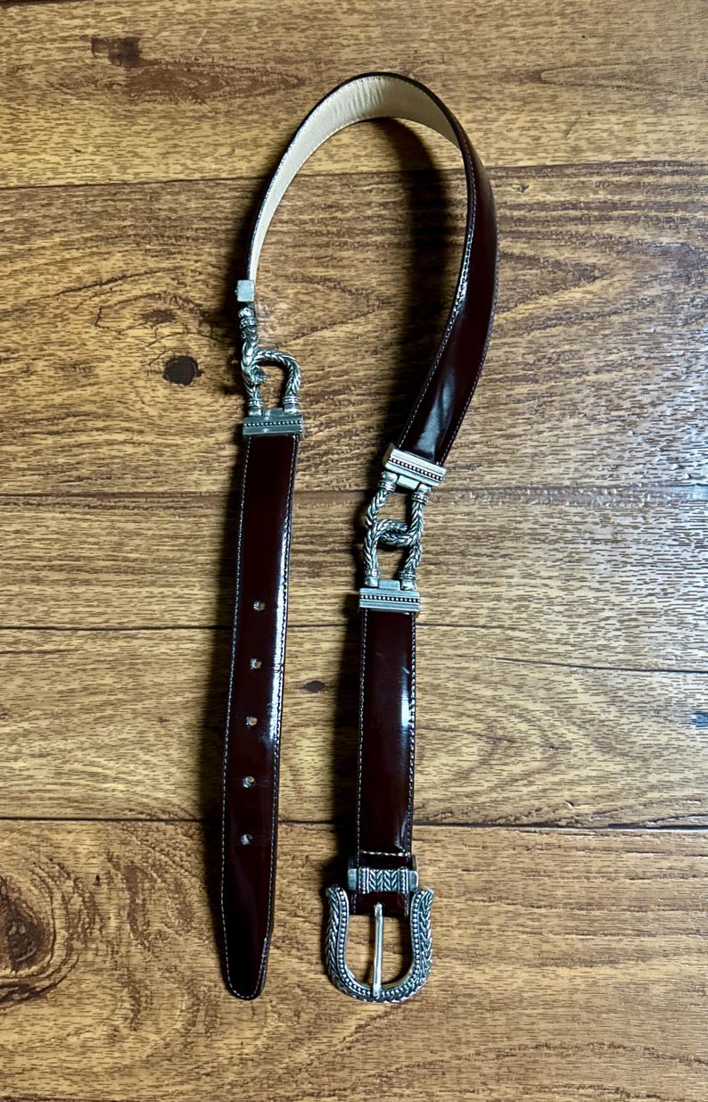 Puede incluir: Un cintur&oacute;n de cuero marr&oacute;n con una hebilla plateada. La hebilla tiene un dise&ntilde;o decorativo con un patr&oacute;n de cuerda.