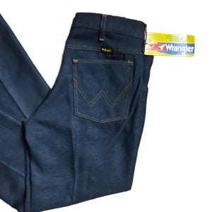 Może przedstawiać: Ciemnoniebieskie dżinsy Wrangler z klasycznym pięciokieszeniowym designem. Tylna kieszeń ma charakterystyczne przeszycia "W" w kontrastującym beżowym odcieniu. Do dżins&oacute;w dołączona jest ż&oacute;łto-czerwona metka Wrangler.