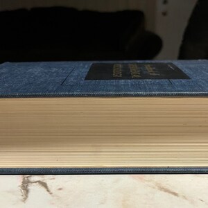 Puede incluir: Un libro de tapa dura azul oscuro con lomo blanco y letras doradas. El libro est&aacute; cerrado y sobre una superficie de m&aacute;rmol.