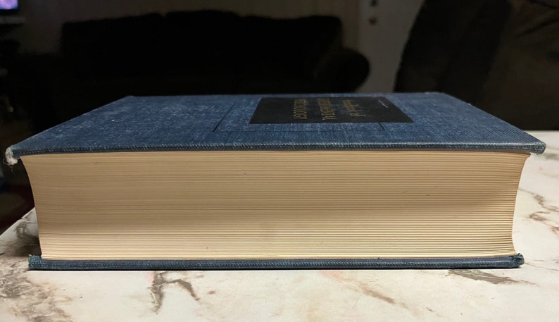 Puede incluir: Un libro de tapa dura azul oscuro con lomo blanco y letras doradas. El libro est&aacute; cerrado y sobre una superficie de m&aacute;rmol.