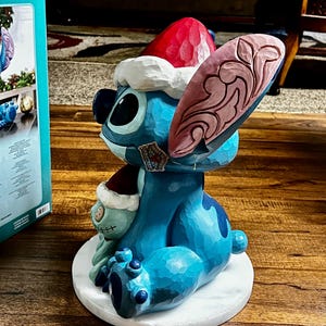 May include: A blue Stitch figurine wearing a red and white Santa hat, with a smaller Stitch figure. The figurine has a carved wood appearance and sits on a white circular base. A Disney tag is visible.