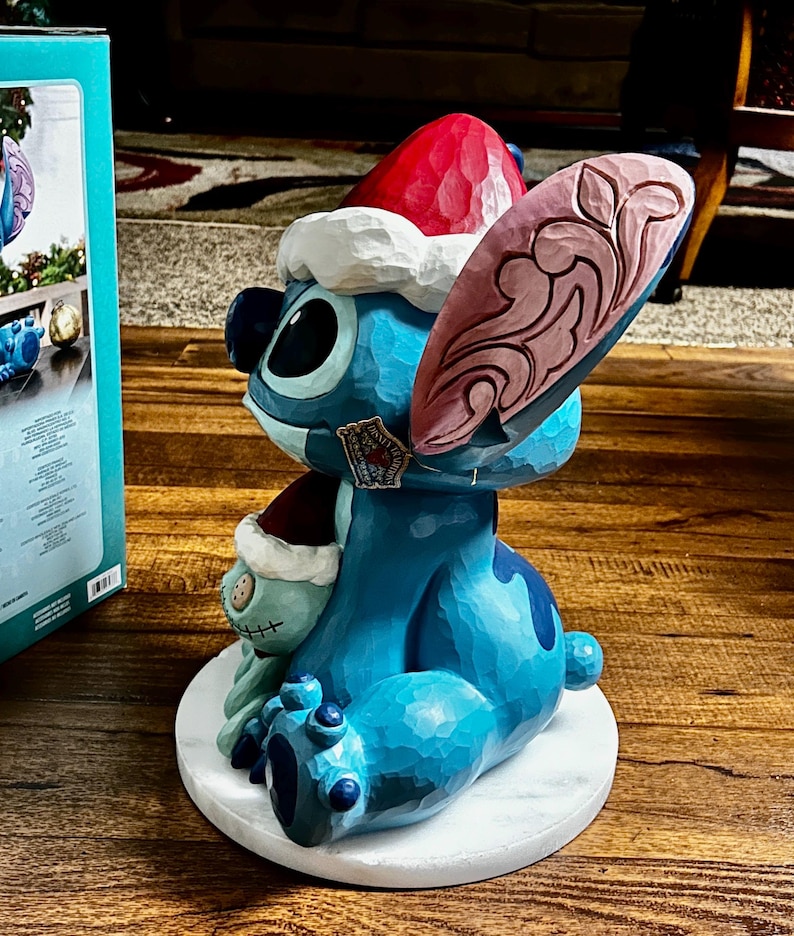 May include: A blue Stitch figurine wearing a red and white Santa hat, with a smaller Stitch figure. The figurine has a carved wood appearance and sits on a white circular base. A Disney tag is visible.