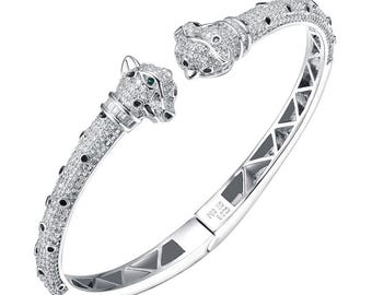 May include: A silver bangle bracelet with two panther heads on the ends. The panther heads are encrusted with clear and green gemstones. The bracelet is engraved with the text "925 S925".