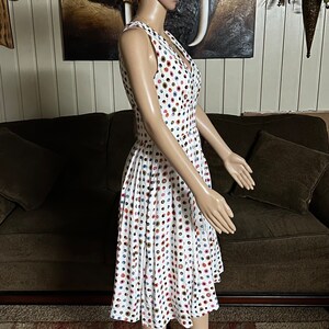 May include: White sleeveless dress with a colorful polka dot pattern. The dress features a V-neckline and a button-down front. The skirt is pleated, and the dress falls to the mid-calf. The dress is displayed on a mannequin.