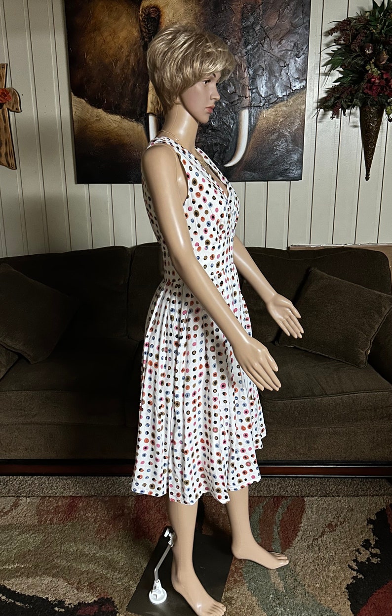 May include: White sleeveless dress with a colorful polka dot pattern. The dress features a V-neckline and a button-down front. The skirt is pleated, and the dress falls to the mid-calf. The dress is displayed on a mannequin.