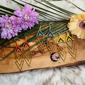 May include: Four pairs of geometric triangle earrings with beaded details. The earrings are in orange, yellow, blue, and green colours. The earrings are on a wooden surface with flowers.