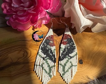 May include: Handmade beaded earrings with a unique wing design. The earrings feature a pattern of white, black, red, and green beads, with a fringe at the bottom. The earrings are displayed on a wooden surface with pink and white flowers in the background.