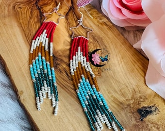 May include: A pair of beaded earrings with a star-shaped top and long fringe. The earrings feature red, white, turquoise, and brown beads. The earrings are displayed on a wooden surface with pink and purple flowers in the background.