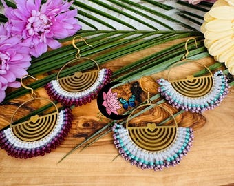 May include: Three pairs of gold hoop earrings with a heart design and a beaded fringe. The fringe is made of white, purple, and teal beads. The earrings are displayed on a wooden surface with green leaves and pink flowers.