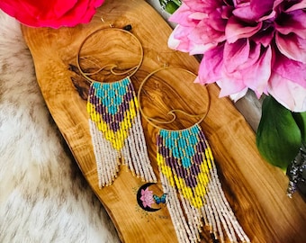 May include: Gold hoop earrings with beaded fringe in turquoise, yellow, purple, and white. The earrings are displayed on a wooden board with flowers in the background. The earrings have a wave design.
