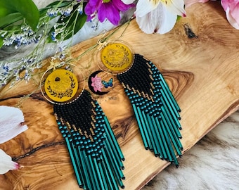 May include: A pair of handmade earrings with a circular design. The earrings feature a black beaded top with teal fringe. The circular design is yellow with a gold-colored rim. The earrings are displayed on a wooden surface with flowers.