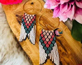 May include: A pair of gold-toned hoop earrings with a wave design, featuring beaded fringe in a geometric pattern of black, white, pink, and teal. The earrings are displayed on a wooden surface with pink and red flowers.